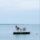 A person sits alone on a small dock with two empty chairs, surrounded by calm blue water under a cloudy sky, evoking the quiet intimacy often found in dreamy tablescapes inspired by An Entertaining Story from Rizzoli.