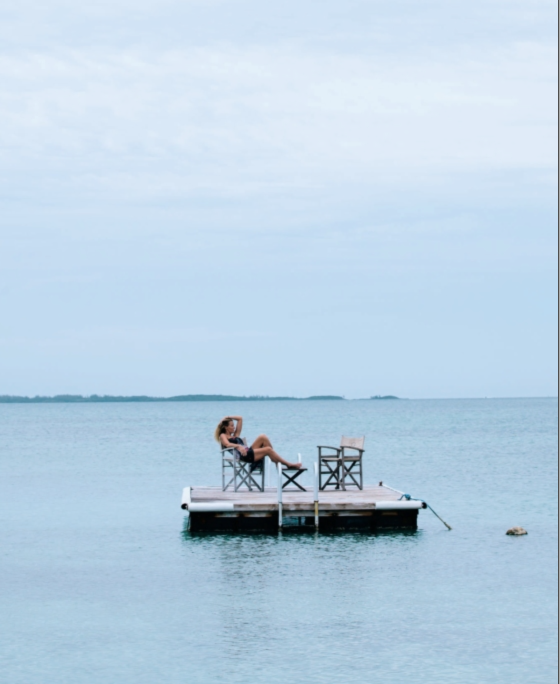 A person sits alone on a small dock with two empty chairs, surrounded by calm blue water under a cloudy sky, evoking the quiet intimacy often found in dreamy tablescapes inspired by An Entertaining Story from Rizzoli.