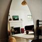 Discover the thoughtfully designed interiors featured in AD At Home: Architectural Digest by Penguin Random House, where a large, irregularly shaped mirror reflects a cozy room adorned with a fireplace, art, small sculptures, a bed, and a window overlooking wooden floors. Natural light streams in to highlight sophisticated design elements that showcase elevated taste and authoritative style.