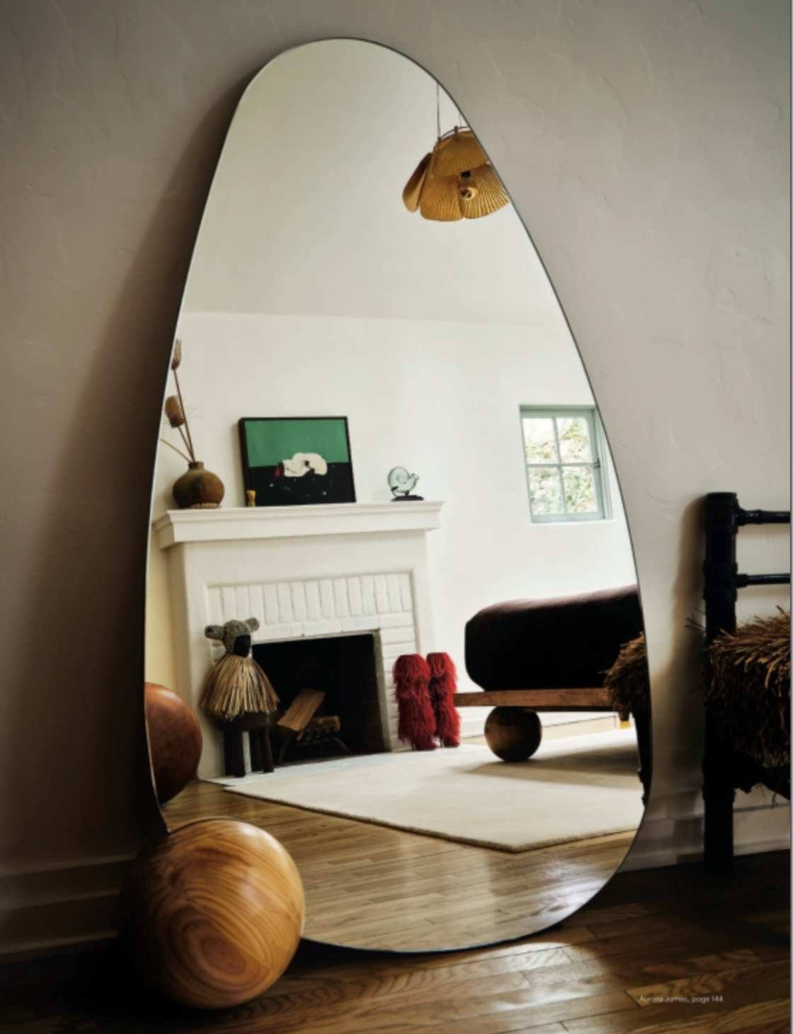 Discover the thoughtfully designed interiors featured in AD At Home: Architectural Digest by Penguin Random House, where a large, irregularly shaped mirror reflects a cozy room adorned with a fireplace, art, small sculptures, a bed, and a window overlooking wooden floors. Natural light streams in to highlight sophisticated design elements that showcase elevated taste and authoritative style.
