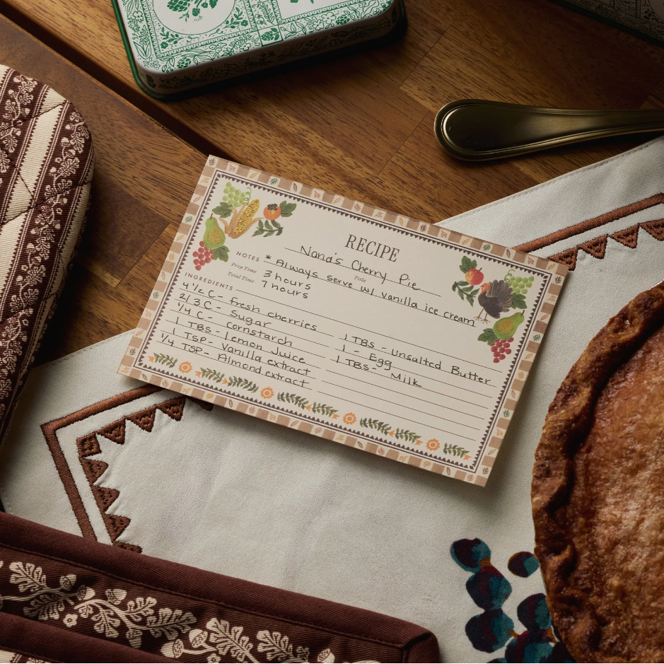 A handwritten cherry pie recipe card sits on a patterned cloth near a baked pie, a brown patterned oven mitt, and a decorative tin on a wooden table—perfect inspiration for your own Rifle Paper Co. Harvest Recipe Cards or special Thanksgiving recipes.