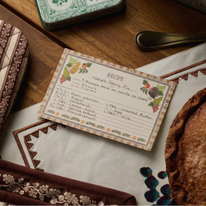 A handwritten cherry pie recipe card sits on a patterned cloth near a baked pie, a brown patterned oven mitt, and a decorative tin on a wooden table—perfect inspiration for your own Rifle Paper Co. Harvest Recipe Cards or special Thanksgiving recipes.