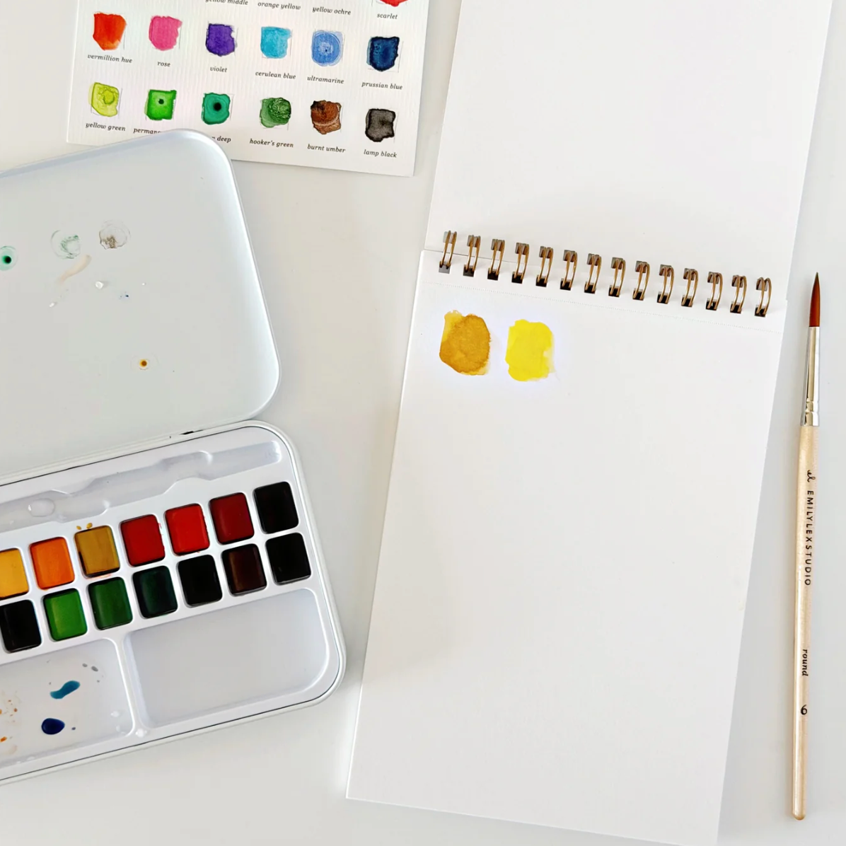 Spiral Watercolor Paper Pad with two yellow paint swatches, a metal palette of watercolor pans, a color reference sheet, and a round paintbrush.