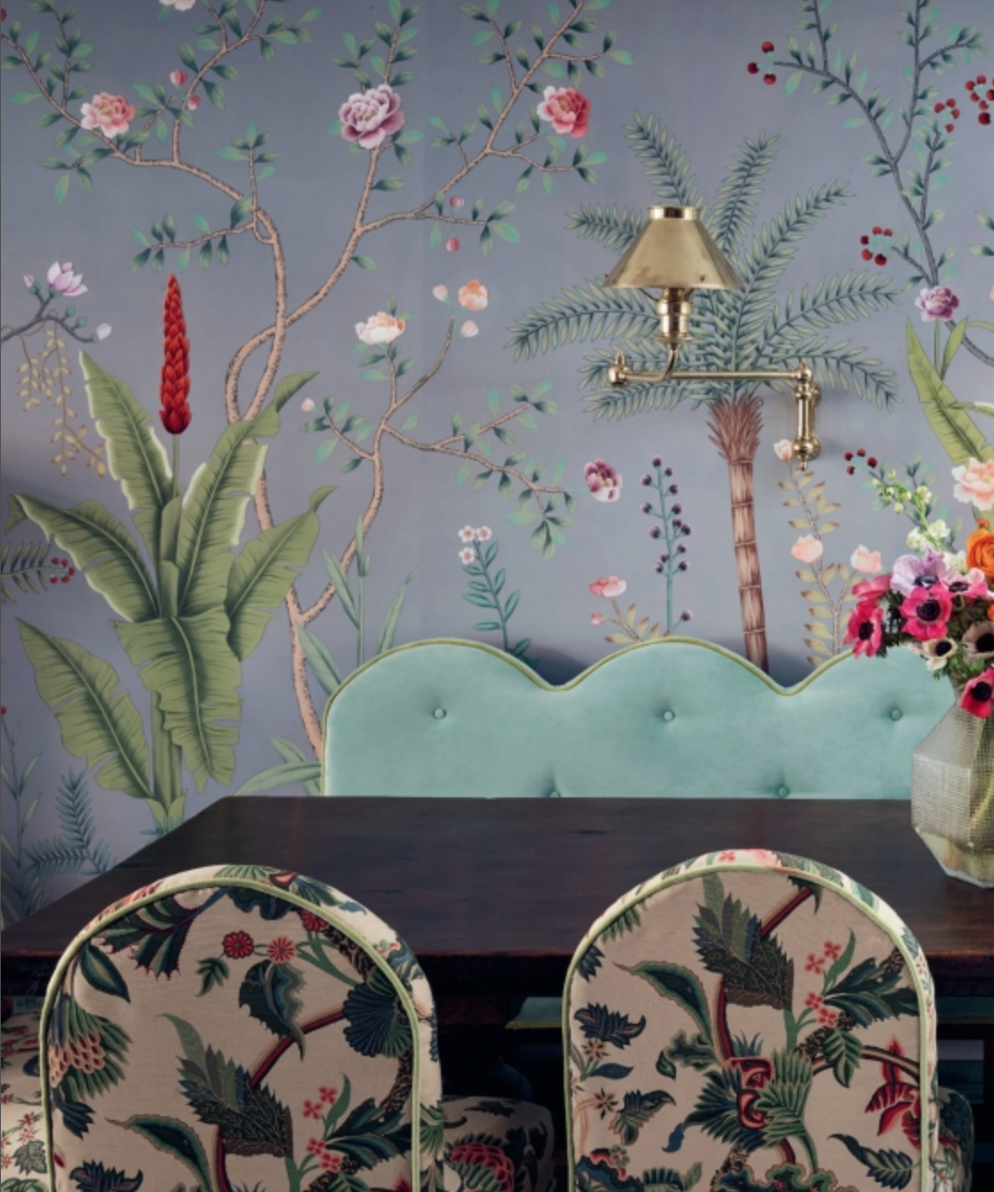 Dining table with floral chairs and a pale blue scalloped bench against de Gournay: Hand-Painted Interiors wallpaper featuring trees, flowers, and a brass wall lamp.