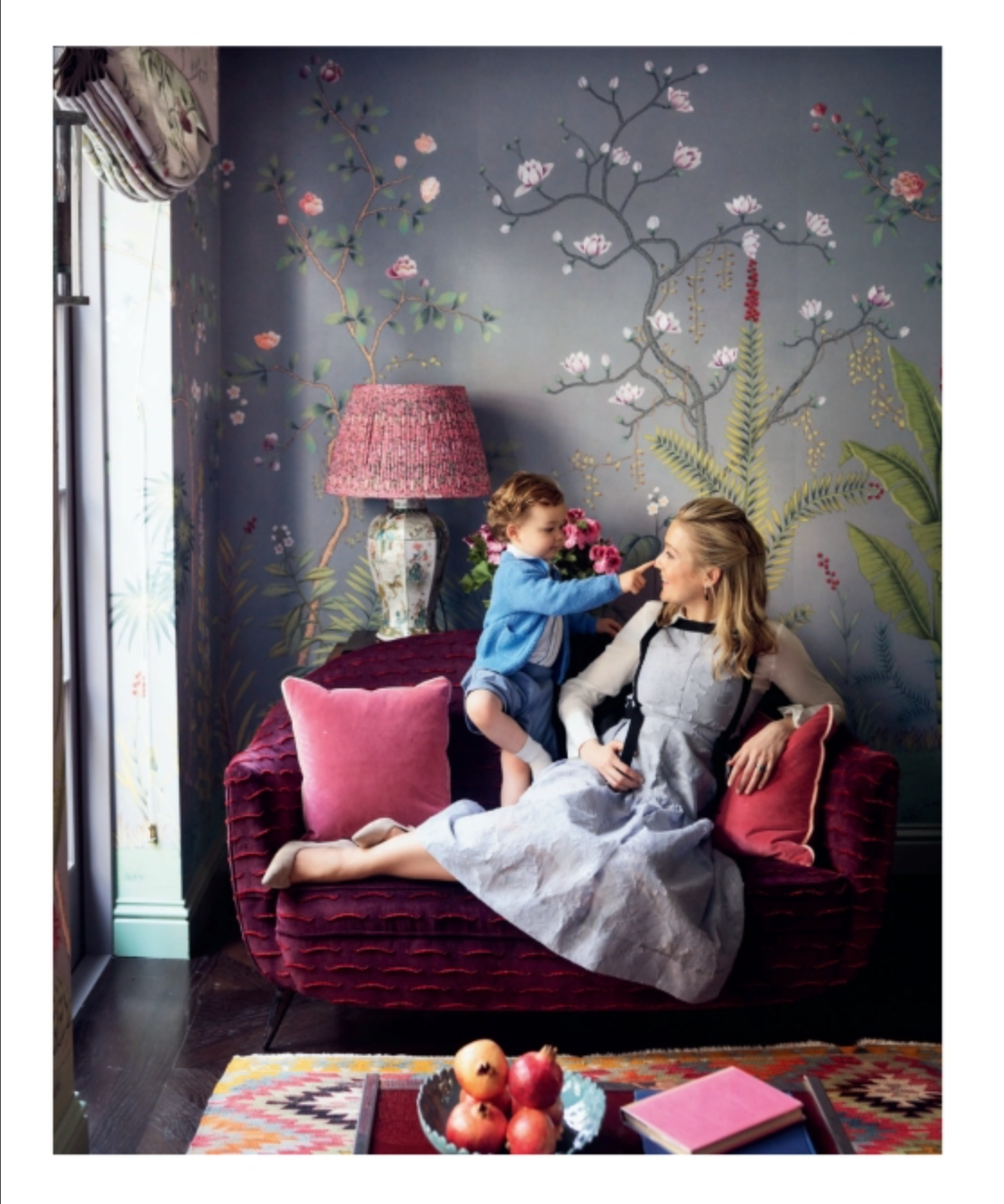 A woman and a young child sit on a velvet sofa against a wall decorated with de Gournay: Hand-Painted Interiors chinoiserie wallpaper.