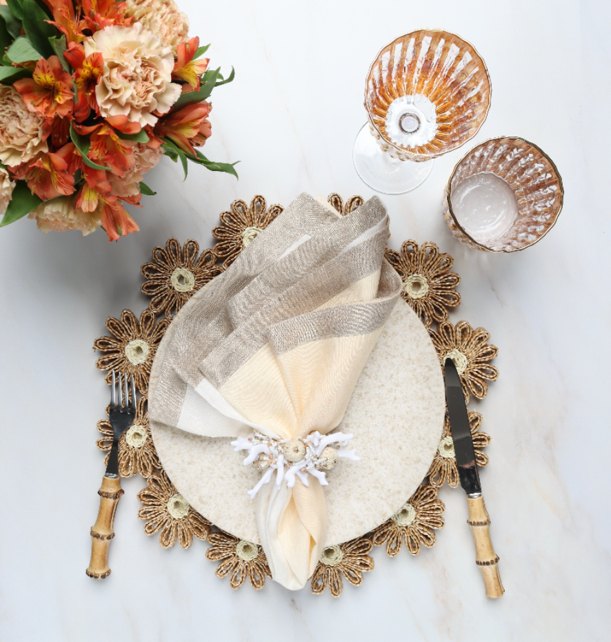 A table setting featuring the Kim Seybert Daisy Placemat, beige napkin with ring, two glasses, bamboo-handled cutlery, and orange flowers on a white surface—ideal for stylish outdoor gatherings.