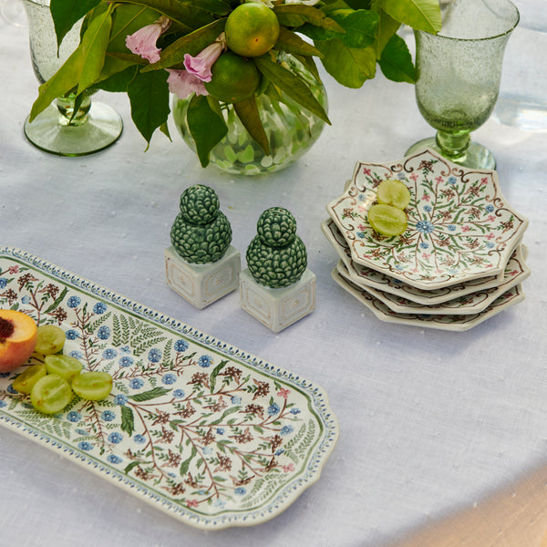 A table set with patterned ceramic dishes, green glasses, a peach, grapes, and Juliska's Berry & Thread Topiary Salt and Pepper Set evokes formal garden charm, while a vase of greenery adds an elegant finishing touch.
