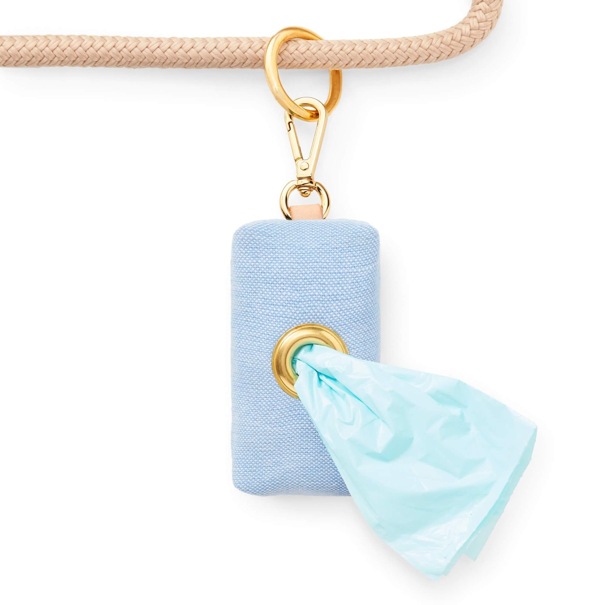 The Foggy Dog’s Waste Bag Dispenser in Blue Chambray features a gold clip and attaches seamlessly to your beige leash, dispensing light blue waste bags through a sturdy metal ring for a chic and stylish pet accessory.
