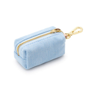 The Foggy Dog Waste Bag Dispenser in Blue Chambray is a chic, light blue rectangular fabric pouch featuring a gold zipper and an attached gold metal clip; shown on a white background, this elegant accessory keeps pet waste bags stylishly at hand.