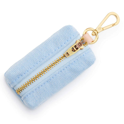 The Foggy Dog’s Waste Bag Dispenser in Blue Chambray features a light blue fabric pouch with a gold zipper and clip, shown against a white background. This chic accessory keeps pet waste bags handy, adding both style and function to your walks.