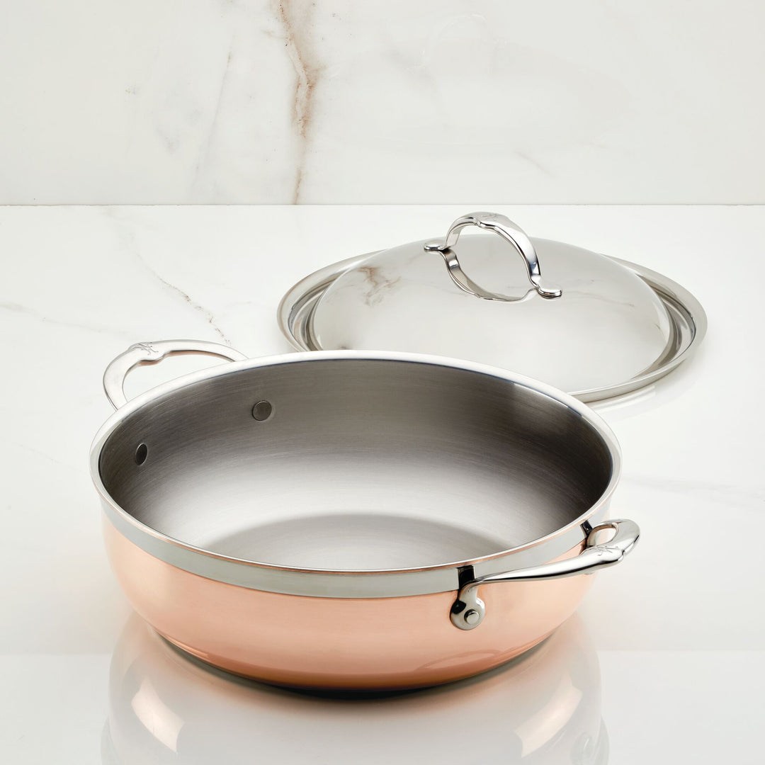 A Hestan CopperBond Copper Induction Dutch Oven, 5qt, with dual handles is shown on a white marble surface with its matching stainless steel lid resting behind it.