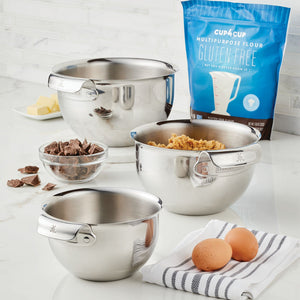 A Hestan Provisions Stainless Steel Mixing Bowl 3-Piece Set, chocolate pieces, brown sugar, butter, eggs, and a bag of gluten-free flour are arranged on a kitchen counter.