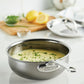 A Hestan NanoBond Titanium Stainless Steel Saucier Pan, 2qt, sits on the counter filled with creamy sauce and garnished with chopped herbs, surrounded by a lid, lemon, and extra herbs in the background.