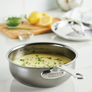 A Hestan NanoBond Titanium Stainless Steel Saucier Pan, 2qt, sits on the counter filled with creamy sauce and garnished with chopped herbs, surrounded by a lid, lemon, and extra herbs in the background.