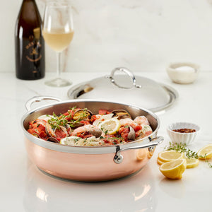 A Hestan CopperBond Copper Induction Dutch Oven, 5qt, filled with seafood and lemon slices sits on a white counter, surrounded by lemon wedges, seasonings, a wine glass, and a bottle.