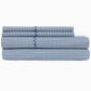 The Cinde Bedding Collection by John Robshaw showcases a neatly folded organic sheet set in Light Indigo with a repeating blue leaf print, including two pillowcases and two larger sheets stacked together.