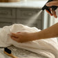A hand sprays L’Avant Collective Unscented Stain Remover onto a white cloth, with a small brush resting nearby on a marble countertop.