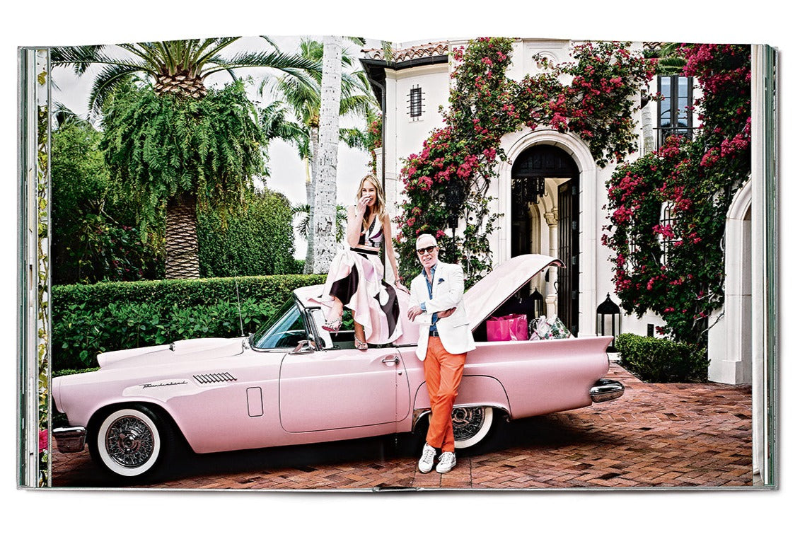 A man stands in front of a pink convertible with the trunk open, positioned beside a woman who is sitting on the car. In the backdrop, an elegant house designed by Martyn Lawrence Bullard and lush greenery paint a picture of ultimate luxury worthy of celebrity homes, featuring Star Style: Interiors of Martyn Lawrence Bullard by Vendome.