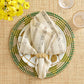 A Stria Ivory & Black Napkin set on a white plate with gold leaf patterns and a woven green and natural-colored charger.