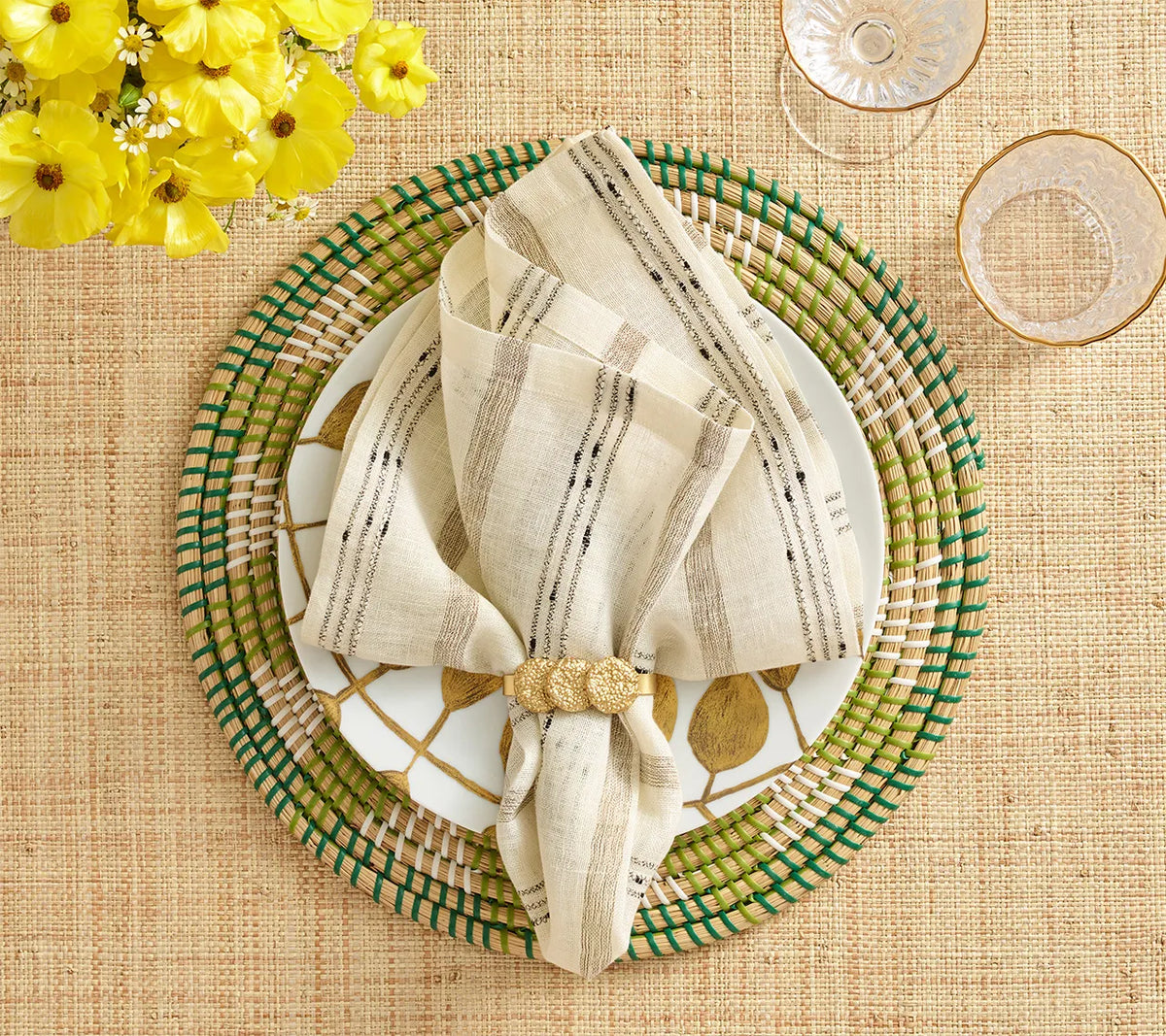 A Stria Ivory & Black Napkin set on a white plate with gold leaf patterns and a woven green and natural-colored charger.