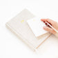 A hand holds a pen over a Gold Initial Mini Notepad resting on a larger fabric-covered journal with gold initials.