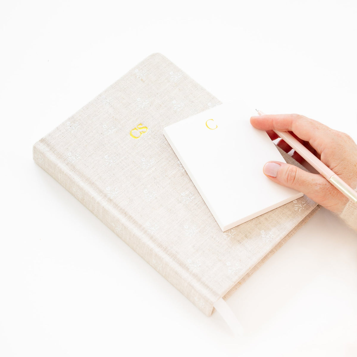 A hand holds a pen over a Gold Initial Mini Notepad resting on a larger fabric-covered journal with gold initials.