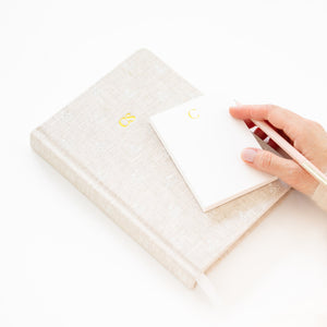 A hand holds a pen over a Gold Initial Mini Notepad resting on a larger fabric-covered journal with gold initials.