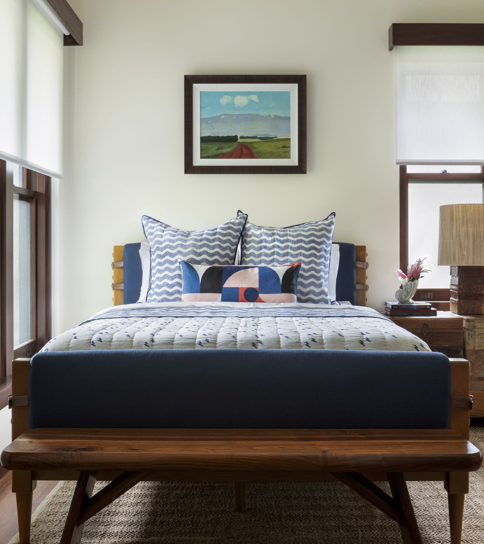 A neatly made bed features Averylily's Surfer Bedding Collection in blue and white, with patterned pillows and a geometric cushion, set below a landscape painting. A wooden bench sits at the foot of the bed.