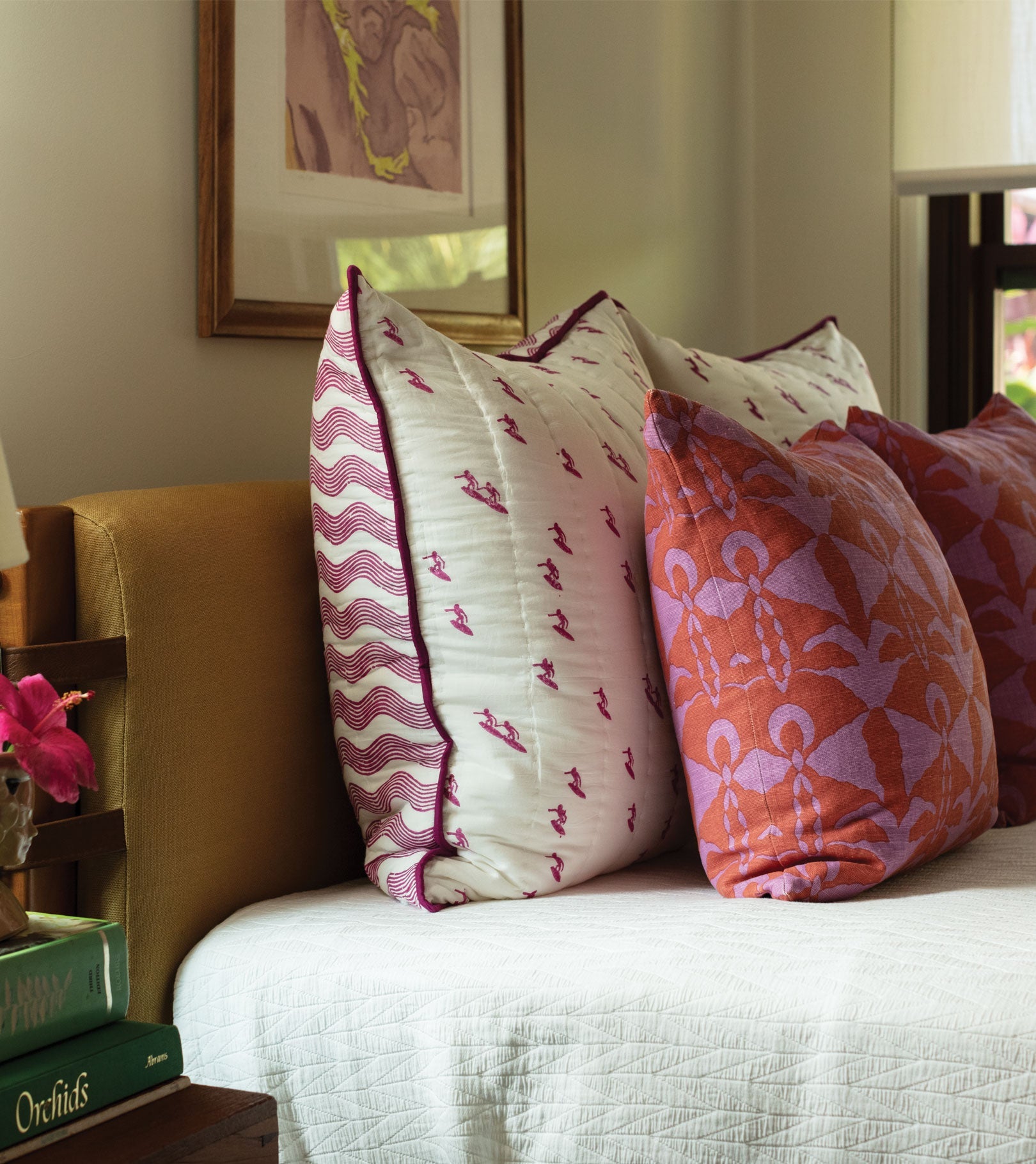 A bed styled with the Averylily Surfer Bedding Collection features white and purple-patterned pillows, orange and purple accents, a white cotton voile bedspread, and framed artwork above.