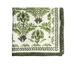 A folded white tablecloth from the Kim Seybert Botanica Tablecloth Collection features a green floral and leaf print, bordered by a green leafy pattern.