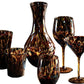 The Juliska Puro Tortoiseshell Drinkware Collection features six handcrafted pieces—small tumblers, wine glasses, a martini glass, and a carafe—elegantly displayed on a white background.