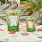 Two Baobab Collection Tomorrowland Candles, featuring intricate designs, are perched on a round stone table, enhancing the poolside area adorned with lounge chairs and palm leaves in the background.