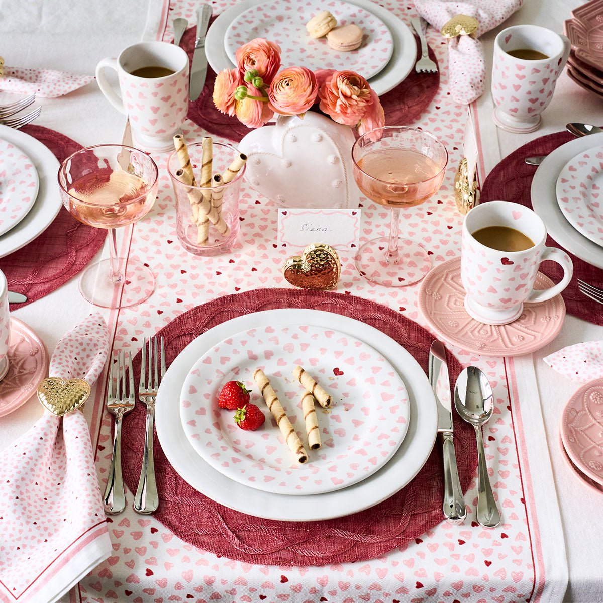 A table set with pink and white heart plates, glasses of rosé, wafer cookies, strawberries, floral décor, and a festive touch from Juliska’s Love You More Table Runner.