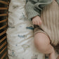 A baby wearing a green sweater and beige knit shorts, crafted from breathable pure materials, lies on a cushion from the Gooselings Transit Treasures Bedding Collection, which is decorated with vintage vehicles including cars, trains, planes, and hot air balloons.