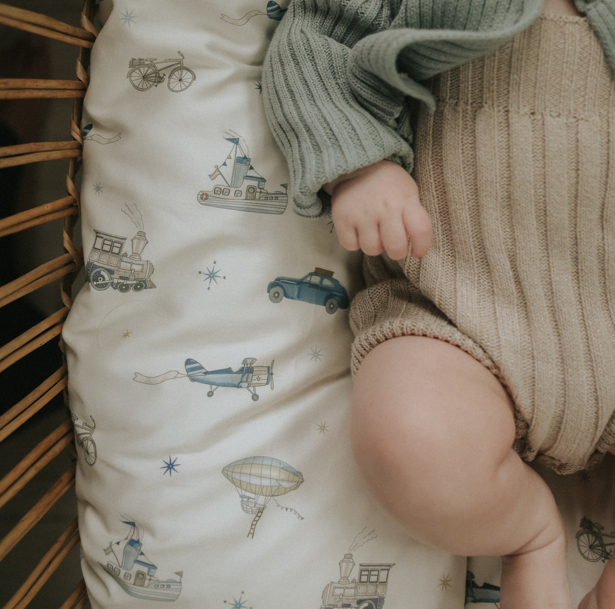 A baby wearing a green sweater and beige knit shorts, crafted from breathable pure materials, lies on a cushion from the Gooselings Transit Treasures Bedding Collection, which is decorated with vintage vehicles including cars, trains, planes, and hot air balloons.