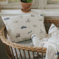 A wicker basket holds the Gooselings Transit Treasures Toddler Pillow and blanket, both featuring vintage vehicles—perfect for bedtime—placed by a window with potted plants.