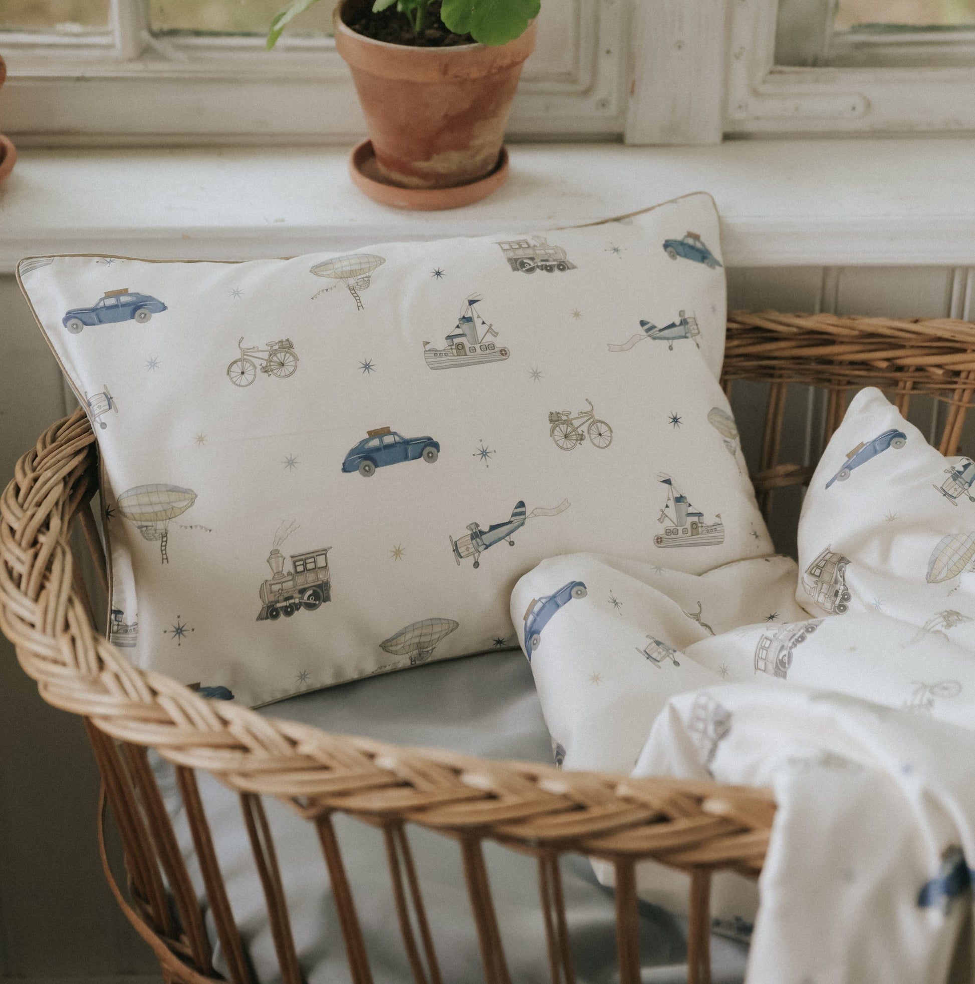 A wicker basket holds the Gooselings Transit Treasures Toddler Pillow and blanket, both featuring vintage vehicles—perfect for bedtime—placed by a window with potted plants.