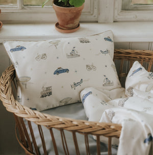 A wicker basket holds the Gooselings Transit Treasures Toddler Pillow and blanket, both featuring vintage vehicles—perfect for bedtime—placed by a window with potted plants.