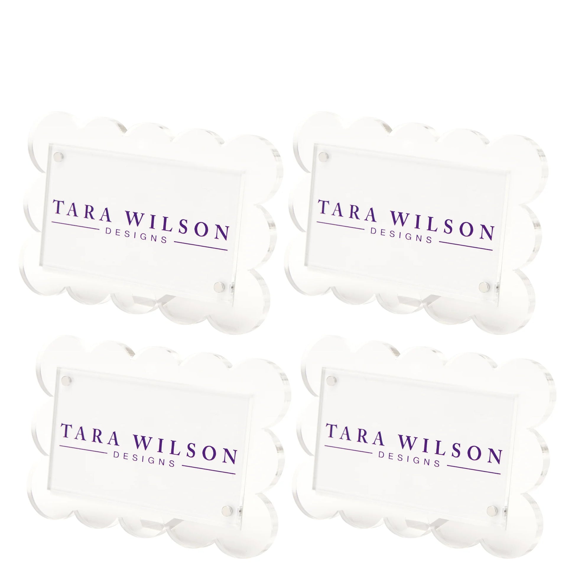 The Scallop Place Card Holder Set by Tara Wilson Designs includes four clear acrylic holders with scalloped edges, each displaying "TARA WILSON DESIGNS" in purple uppercase letters.