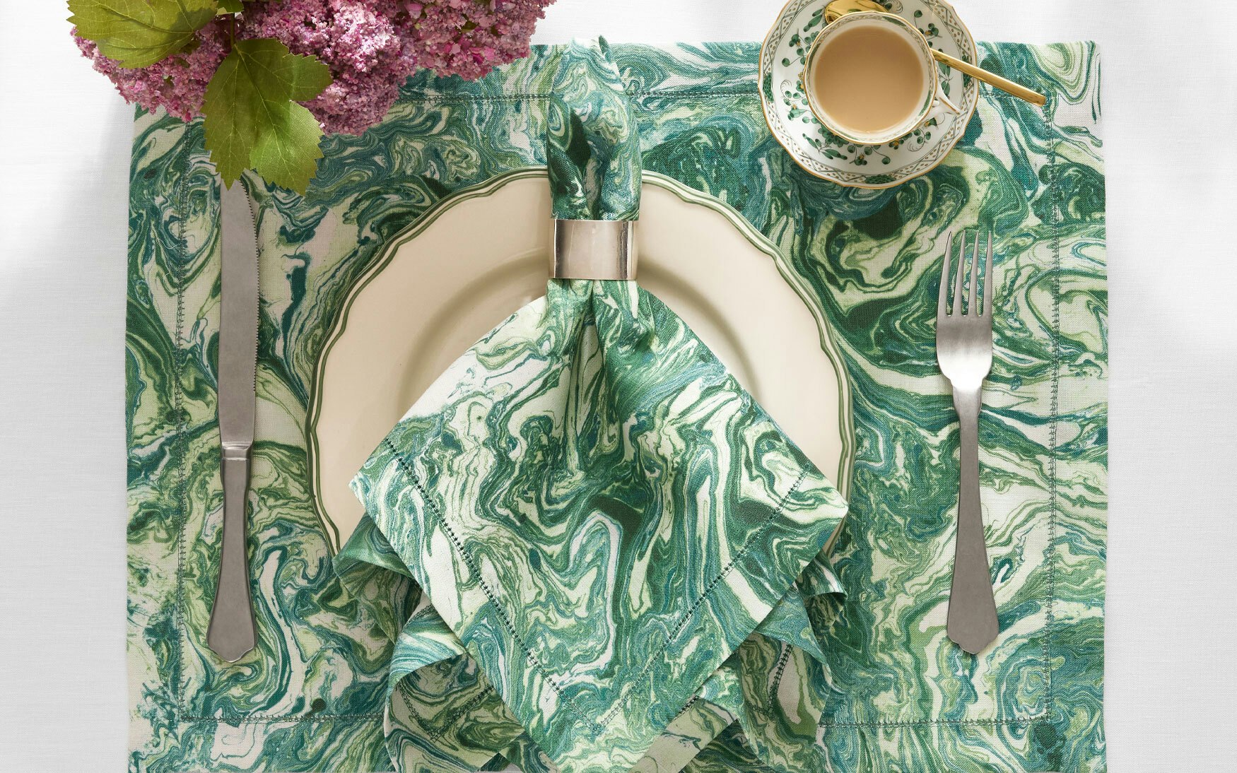 Table setting featuring the Matouk Balthazar Napkin Set in green marble, paired with a white plate, silverware, a cup of coffee, and a bouquet of purple flowers in the corner.