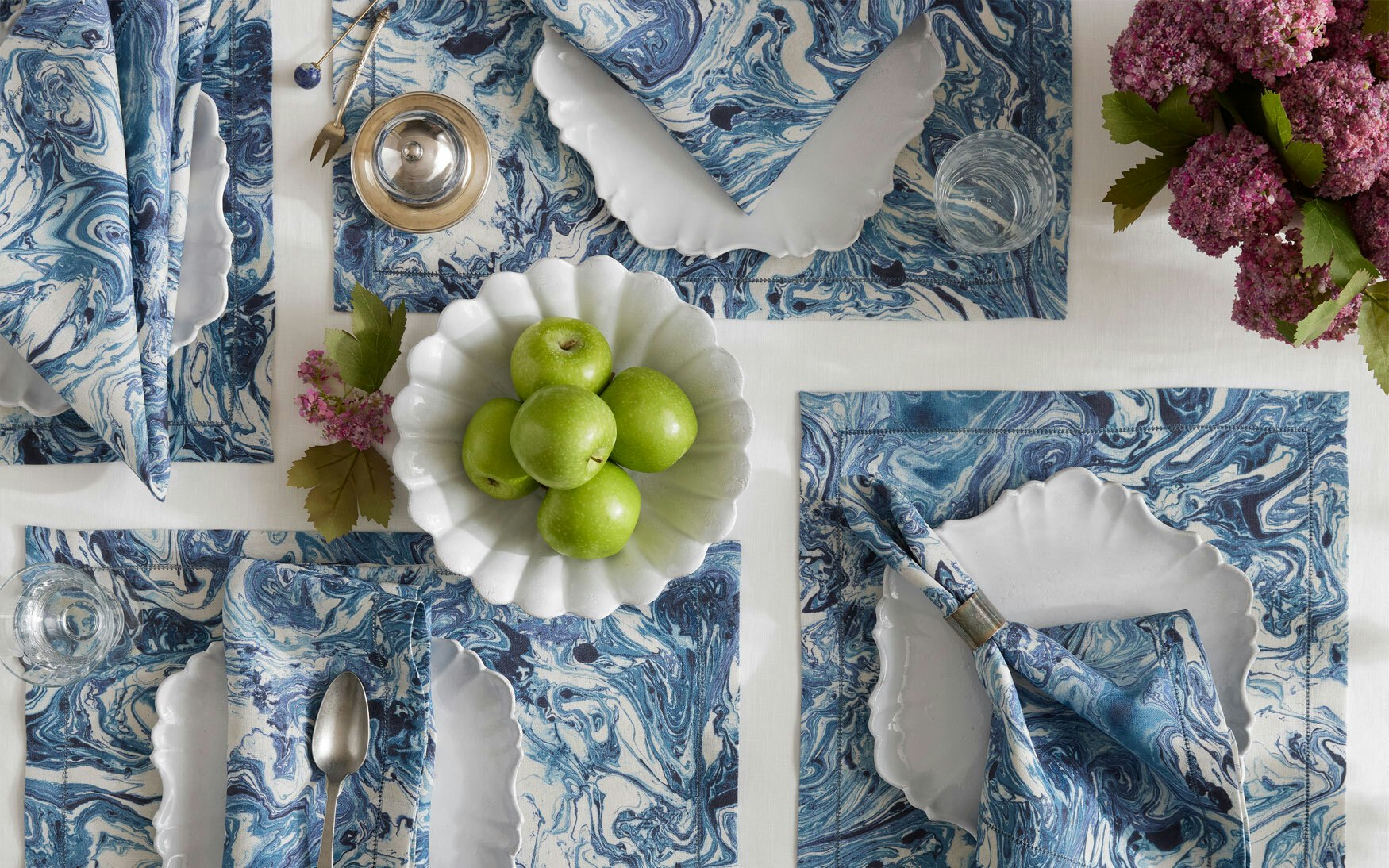 Table set with blue and white marbled table linens, scalloped white plates, silver cutlery, the Balthazar Napkin Set by Matouk, green apples in a bowl, and a bouquet of purple flowers—an elegant touch reminiscent of Matouk style.