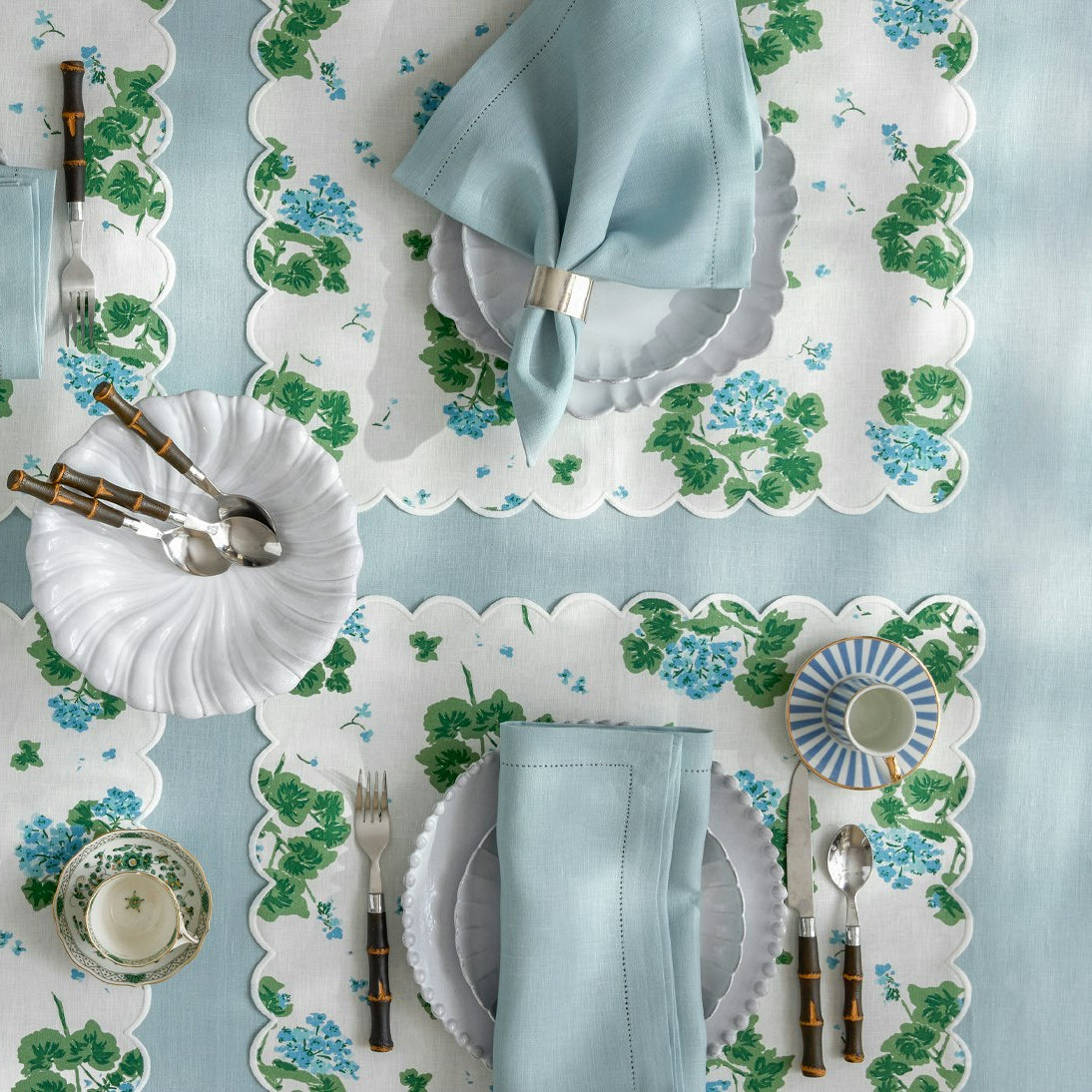 Two place settings with the Matouk Tillett x Matouk Blue Geranium Table Linen Collection, white plates, light blue napkins with silver rings, and shell-shaped dishes create a charming floral table design.