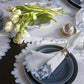 The Wave Tabletop Collection in Hazy Blue by Matouk features blue-rimmed plates, ruffled white napkins, glassware, silverware, and white tulips atop an embroidered linen tabletop and matching ruffled tablecloth.