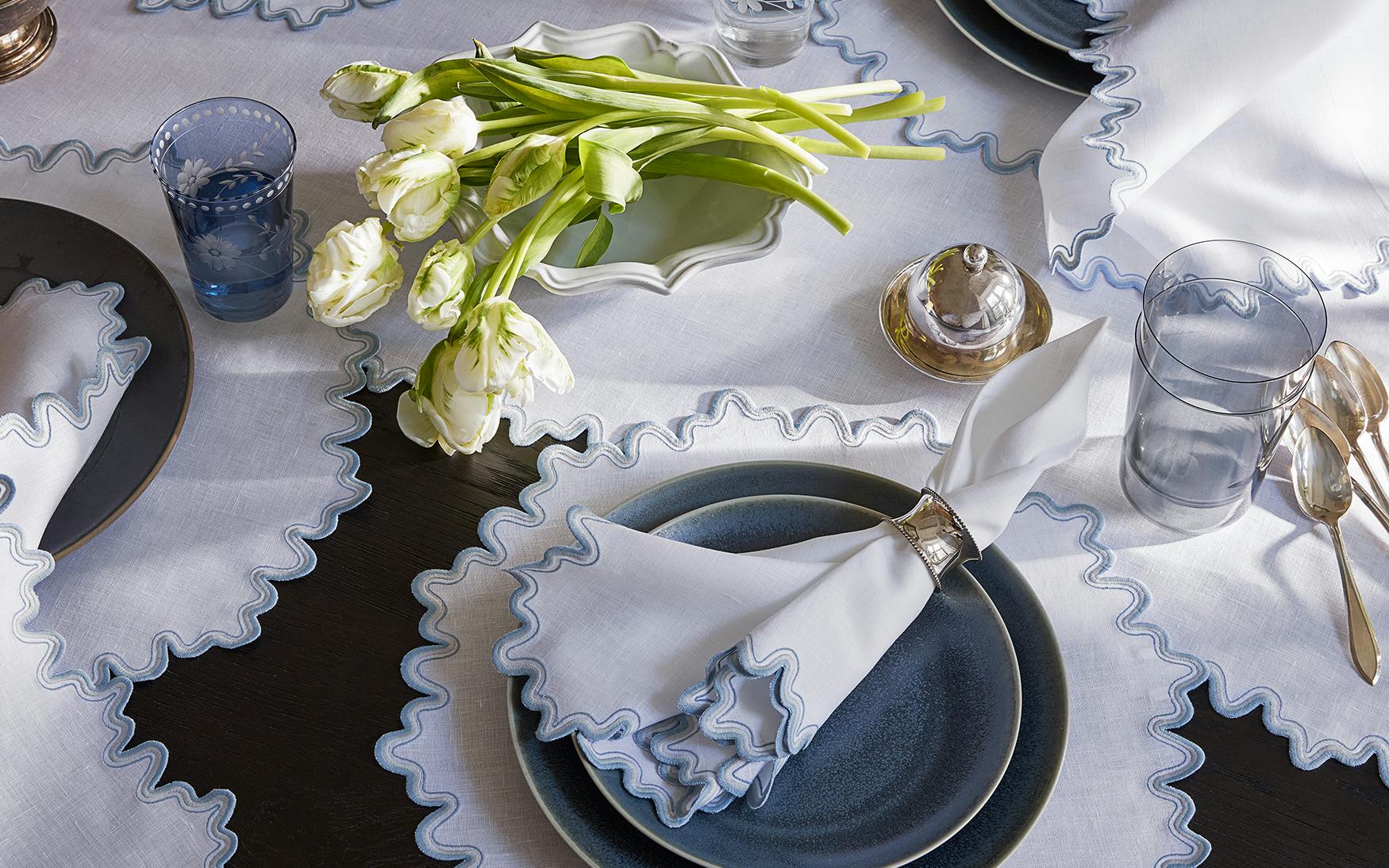 The Wave Tabletop Collection in Hazy Blue by Matouk features blue-rimmed plates, ruffled white napkins, glassware, silverware, and white tulips atop an embroidered linen tabletop and matching ruffled tablecloth.