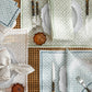 Table set with patterned Laksha Napkin Set by Matouk, scalloped white plates, forks and knives, small brown glass dishes, and green flowers on a woven placemat. The napkins showcase a block printing design inspired by the signature Matouk Schumacher geometric pattern for added visual interest.