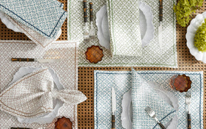 Table set with patterned Laksha Napkin Set by Matouk, scalloped white plates, forks and knives, small brown glass dishes, and green flowers on a woven placemat. The napkins showcase a block printing design inspired by the signature Matouk Schumacher geometric pattern for added visual interest.