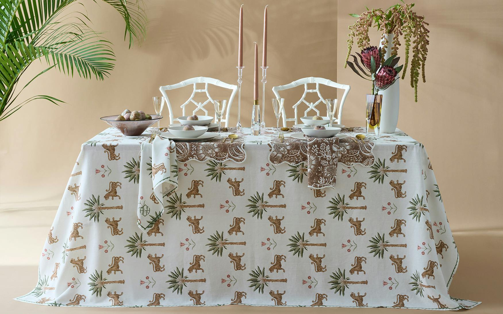 The Tiger Palm Tabletop Collection by Matouk features a set for two with a cheetah-print tablecloth, white chairs, bowls, glasses, candles, and a vase with Ottoman florals beside a potted plant and beige jungle-inspired backdrop.