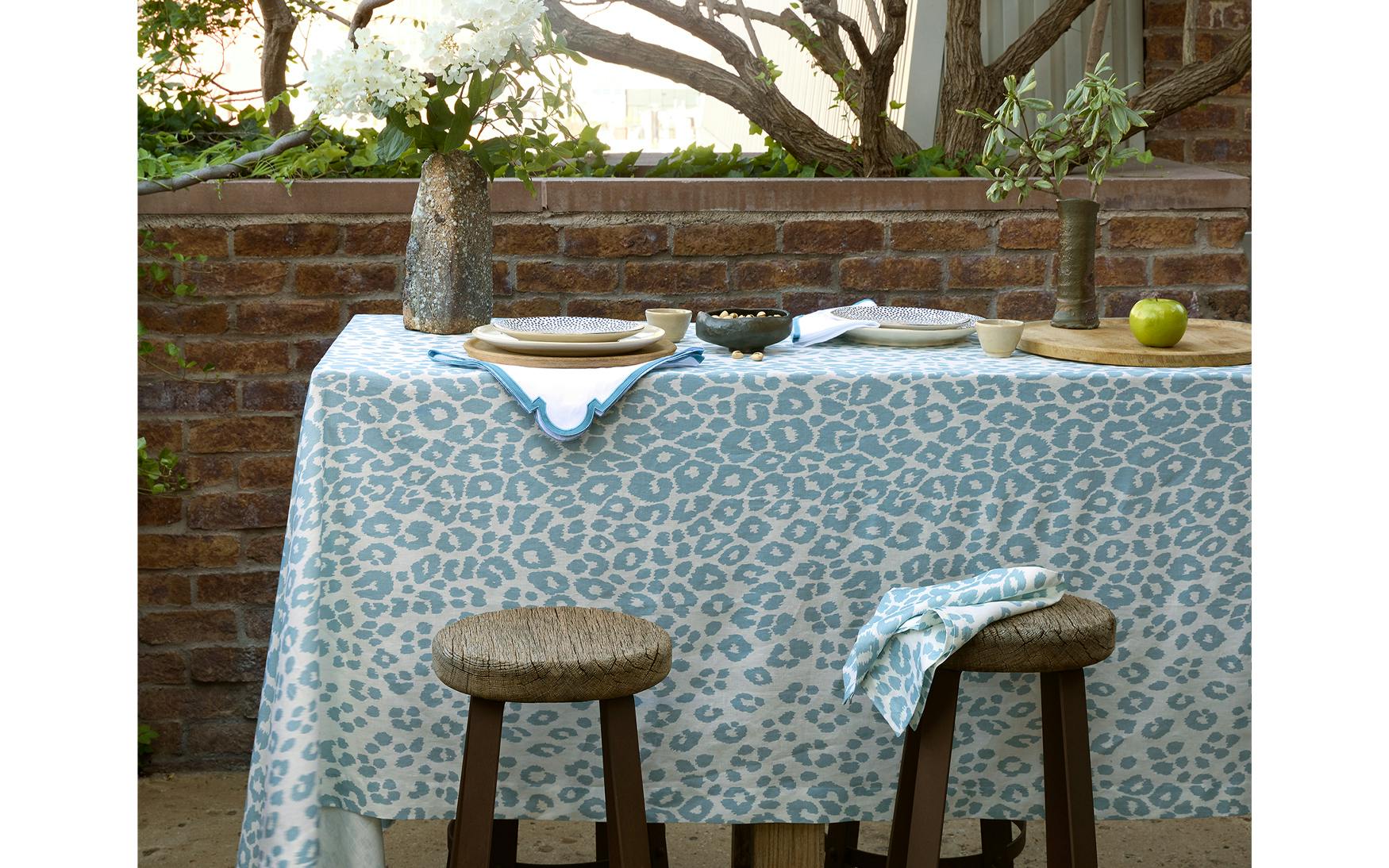 Outdoor table set with dishware and Matouk’s Iconic Leopard Sky Tabletop Collection, featuring a blue tablecloth, two stools, and a brick wall decorated with potted plants—an elegant accent for any classic linen table collection.