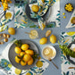 The Matouk Citrus Garden Pool Collection features lemon-patterned napkins, gold cutlery, plates, glasses of lemon water, and pastries arranged on a blue linen tablecloth inspired by Josef Frank’s iconic designs.