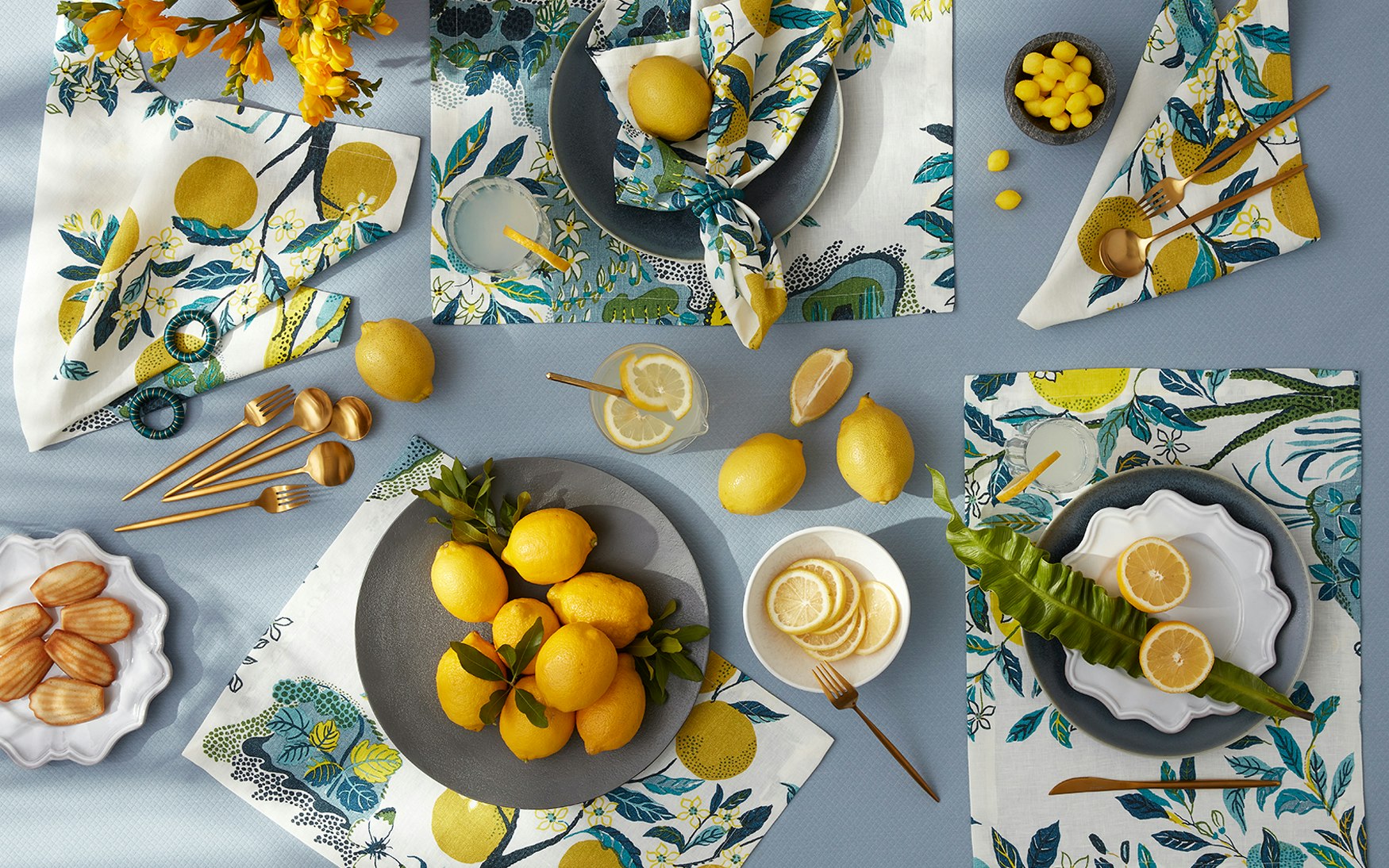The Matouk Citrus Garden Pool Collection features lemon-patterned napkins, gold cutlery, plates, glasses of lemon water, and pastries arranged on a blue linen tablecloth inspired by Josef Frank’s iconic designs.